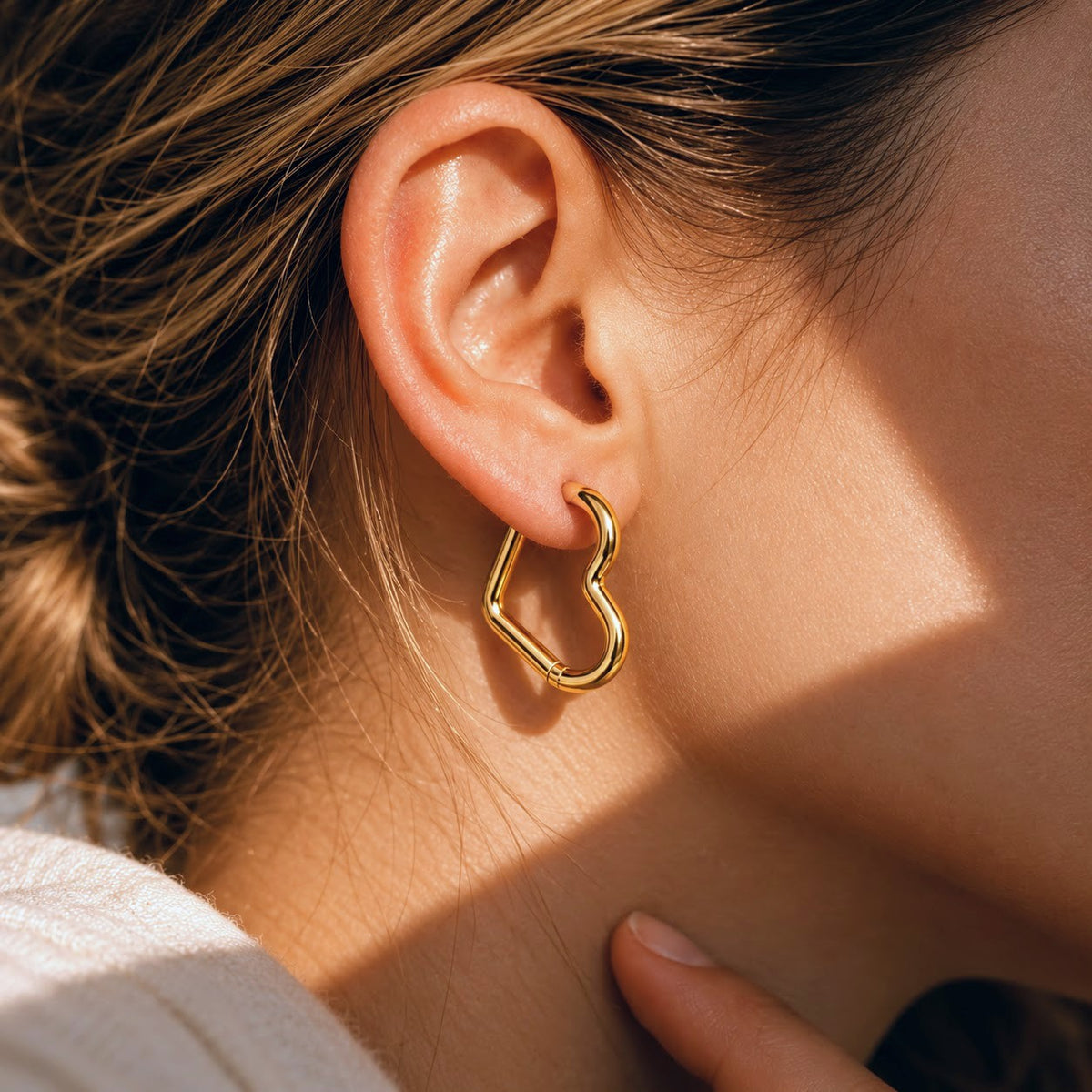 A close-up of a person's ear wearing a polished gold open heart-shaped hoop earring, with warm sunlight casting shadows on the skin.
