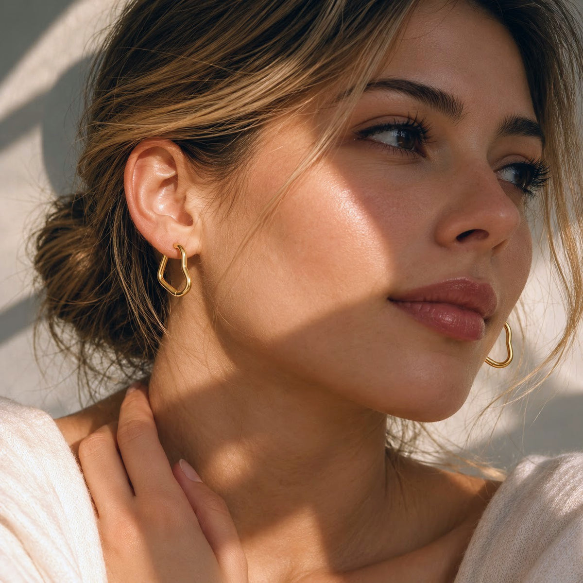 A close-up lifestyle shot of a woman with her hair in a low bun, wearing a small gold huggie hoop earring. The image is characterized by warm, directional sunlight casting soft shadows on her face and neck.