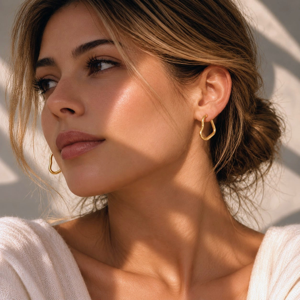 A close-up lifestyle shot of a woman with her hair in a low bun, wearing delicate gold hoop earrings. The image is styled with warm, direct sunlight casting soft shadows on her face and neck, against a neutral light background.