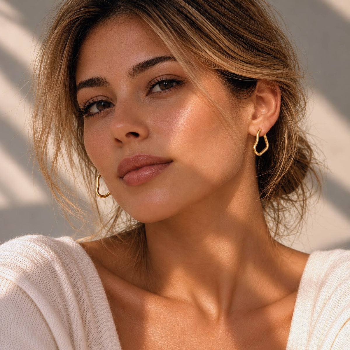 A close-up portrait of a woman wearing delicate gold geometric hoop earrings, styled with a white knit top and natural sunlight casting soft shadows.