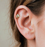A close-up of a person's ear wearing a small gold ear cuff stud decorated with clear crystals, set against a soft beige background.