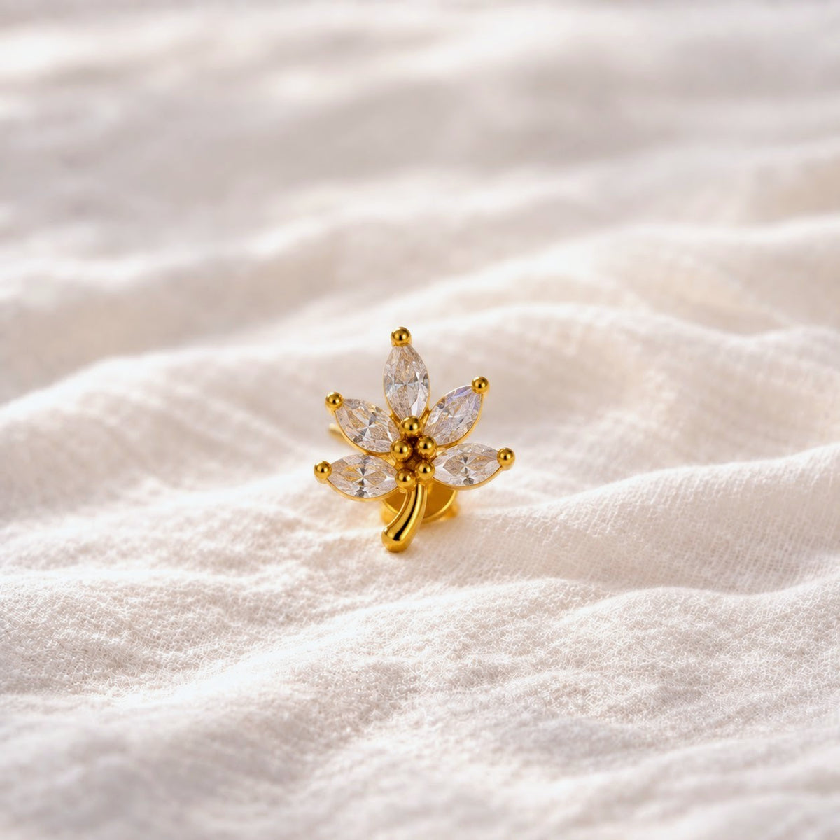A small gold flower-shaped stud earring with marquise-cut clear stones, resting on a soft, textured white fabric background.