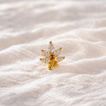 A small gold flower-shaped stud earring with marquise-cut clear stones, resting on a soft, textured white fabric background.