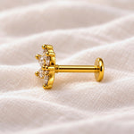 A gold flower-shaped labret stud earring with clear gemstones, resting on a textured beige fabric background.