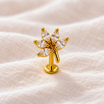 A gold flower-shaped body jewelry piece with marquise-cut clear stones, displayed on a soft beige textured fabric background.