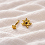 A pair of gold-toned ear piercings, including a flat circular stud and a lotus flower stud with a small dark center stone, displayed on a textured white fabric background.