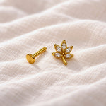A pair of gold-toned ear piercings, including a flat round stud and a lotus flower stud with clear gemstones, resting on a textured beige fabric background.