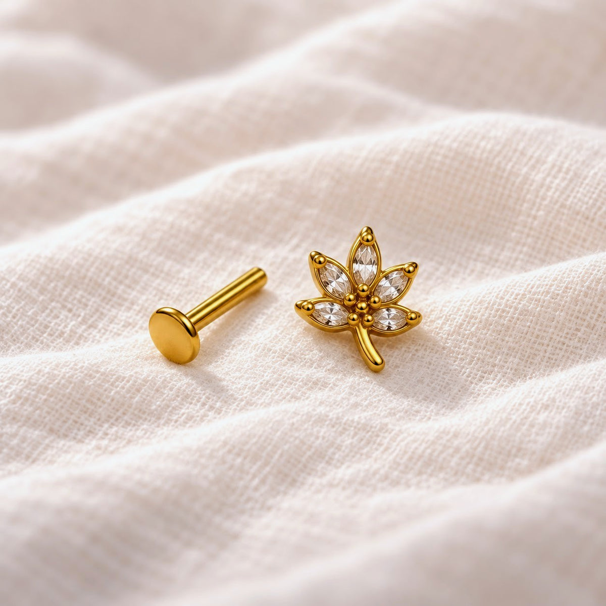 A pair of gold-toned ear piercings, including a flat round stud and a lotus flower stud with clear gemstones, resting on a textured beige fabric background.
