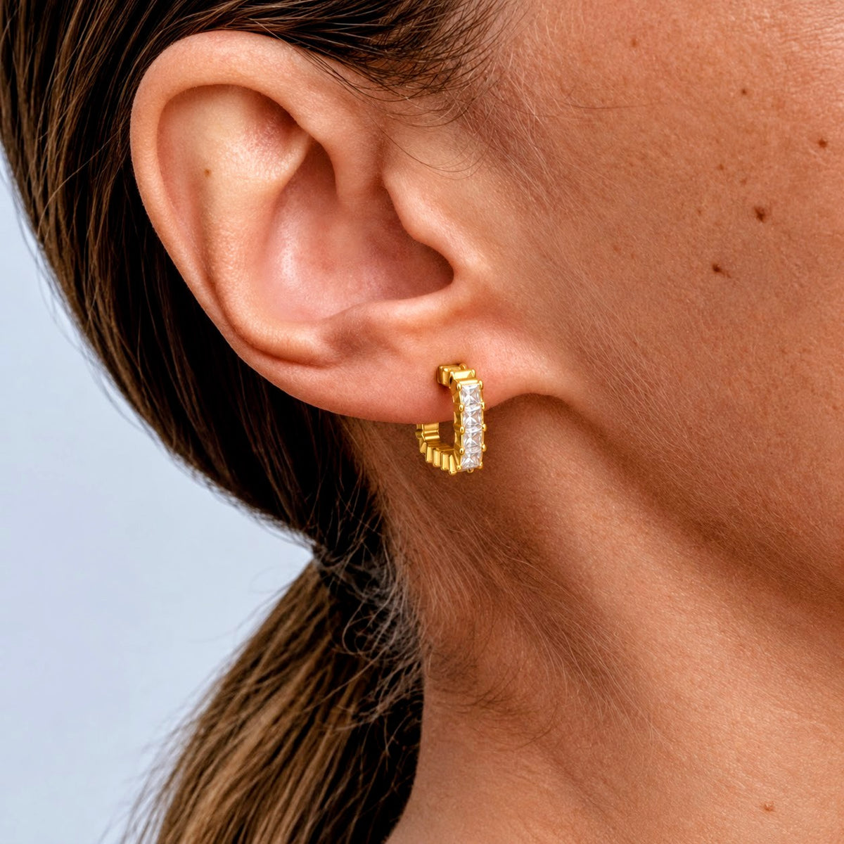 A close-up of a model's ear wearing a small gold huggie hoop earring set with clear pavé stones, against a light gray background.