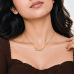A close-up lifestyle shot of a woman wearing a delicate gold chain necklace with a small initial pendant spelling 'LOVE' adorned with tiny clear stones, paired with a brown ribbed off-the-shoulder top.