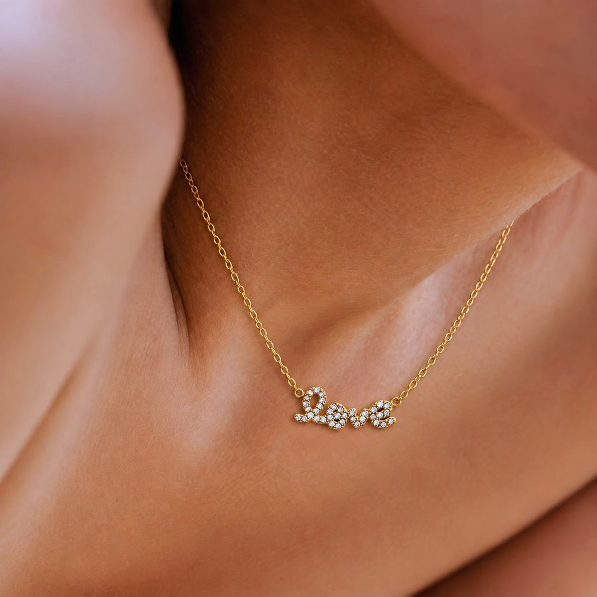 A close-up of a delicate gold chain necklace worn on a person's neck, featuring a small script pendant with clear rhinestones.