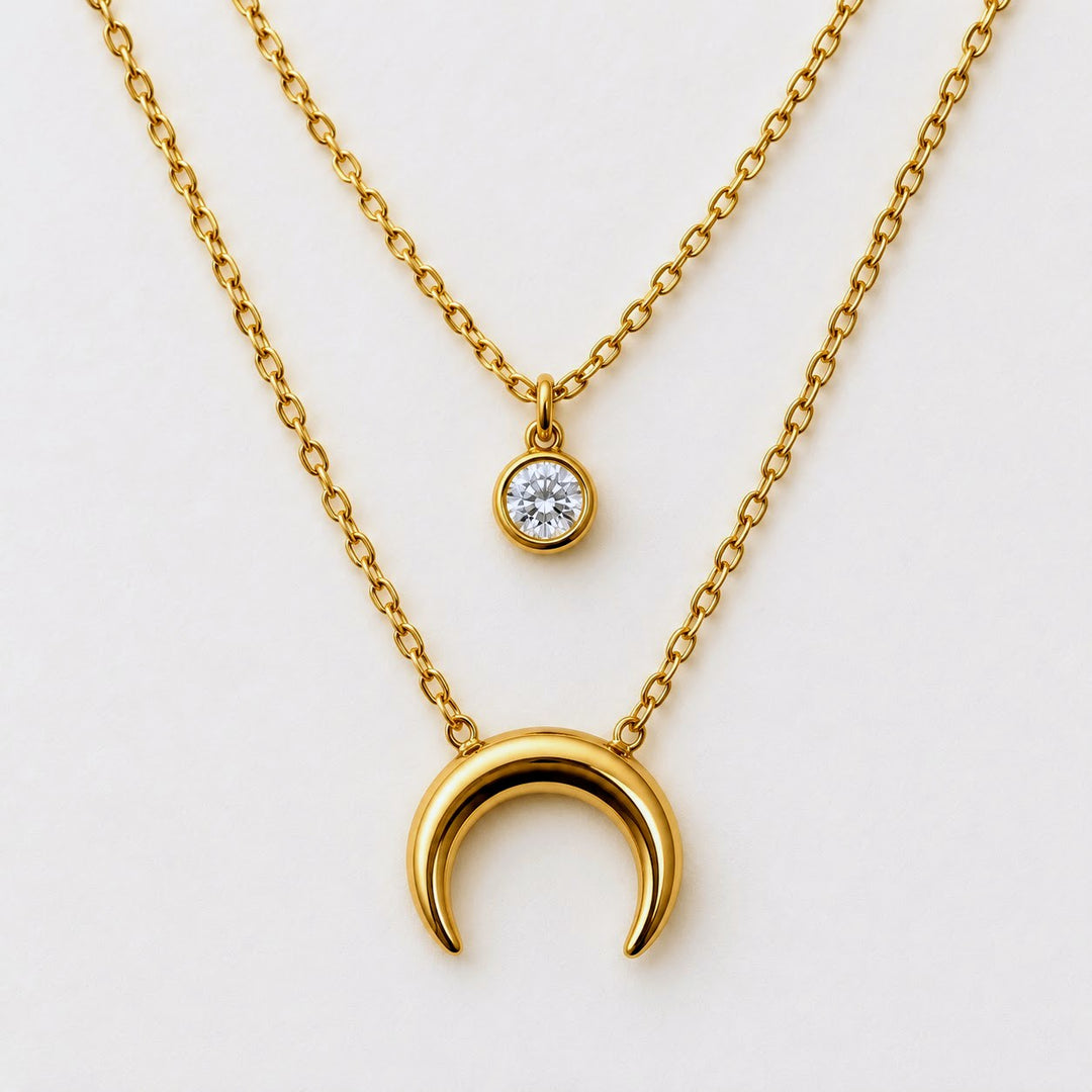 A gold layered necklace set on a clean white background, featuring a larger polished crescent moon pendant and a smaller round bezel-set clear gemstone pendant.