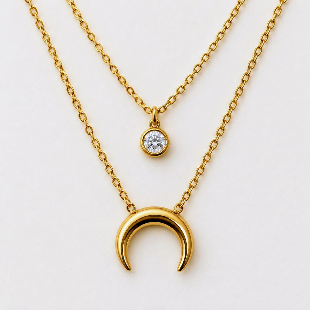 A gold layered necklace set on a clean white background, featuring a larger polished crescent moon pendant and a smaller round bezel-set clear gemstone pendant.