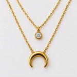 A gold layered necklace set on a clean white background, featuring a larger polished crescent moon pendant and a smaller round bezel-set clear gemstone pendant.