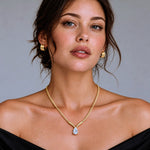 A close-up portrait of a woman wearing a gold chain necklace with a teardrop-shaped clear gemstone pendant, along with matching gold hoop earrings, against a plain gray background.