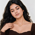 A woman with long, wavy dark hair wearing a delicate gold necklace with the name 'Safia' in cursive script, paired with a brown ribbed off-the-shoulder top against a light gray background.