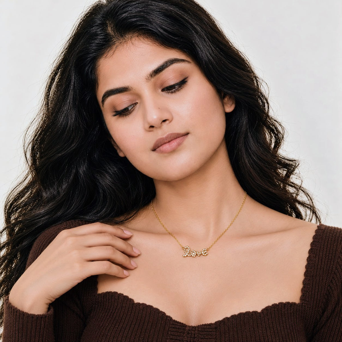 A woman with long, wavy dark hair wearing a delicate gold necklace with the name 'Safia' in cursive script, paired with a brown ribbed off-the-shoulder top against a light gray background.