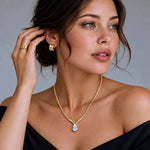 A woman wearing a delicate gold chain necklace with a pear-cut clear gemstone pendant, matching gold hoop earrings, and gold rings, styled with a black off-the-shoulder top against a neutral gray background.