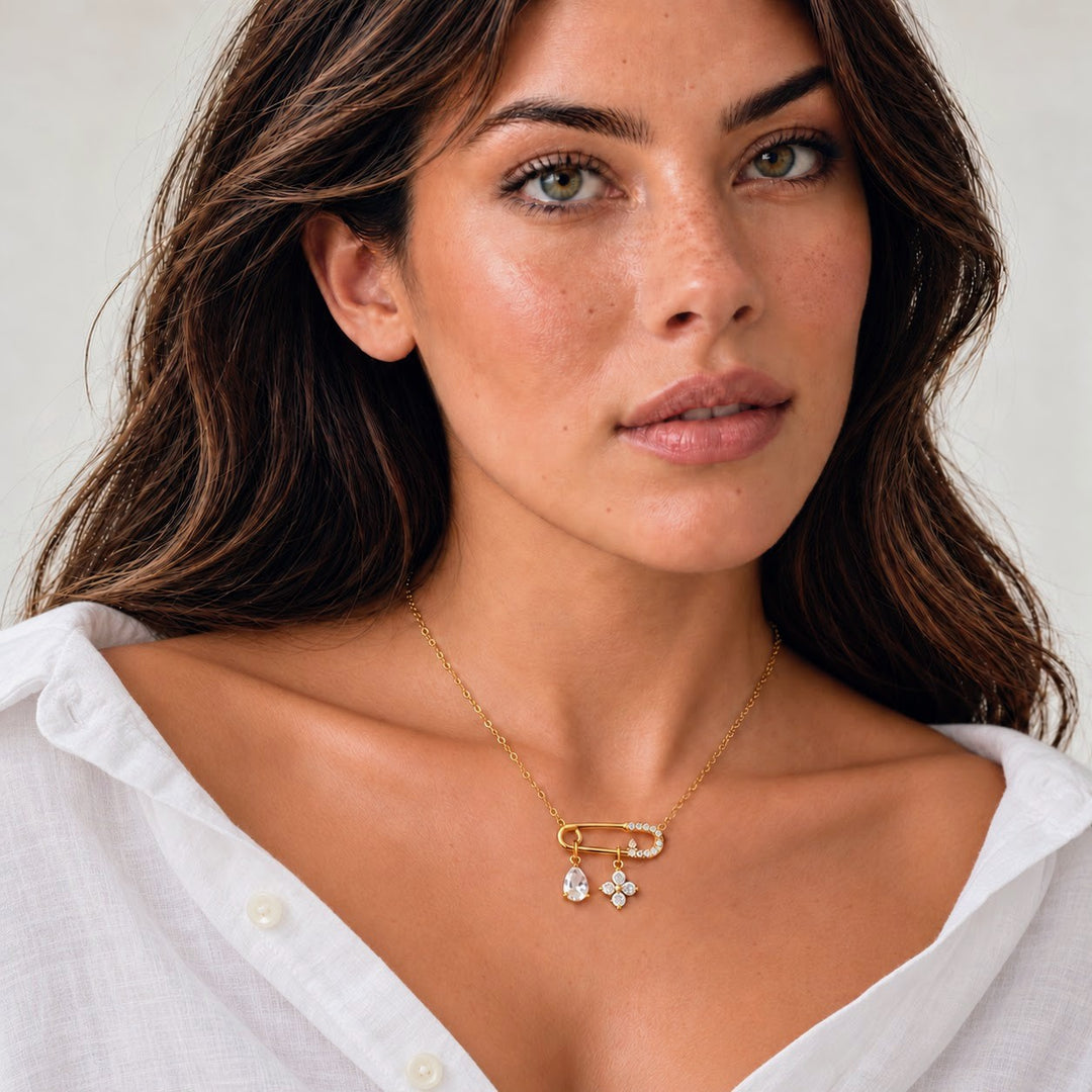 A close-up of a woman wearing a delicate gold necklace with a safety-pin style clasp and two small dangling flower charms, paired with a white collared blouse against a soft light gray background.