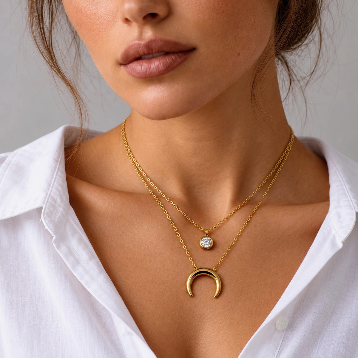 A close-up of a model wearing a double-layered gold necklace. The top layer features a delicate chain with a small round bezel-set clear stone, while the bottom layer has a larger, polished gold crescent moon pendant.