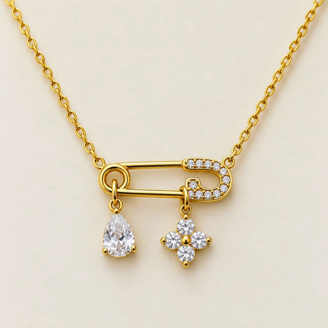 A gold necklace with a safety-pin style pendant adorned with small clear stones, featuring two dangling charms: a pear-shaped clear gemstone and a four-petal flower charm, set against a light beige background.
