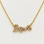 A gold chain necklace with a cursive 'love' pendant paved with small clear stones, shown on a plain light background.