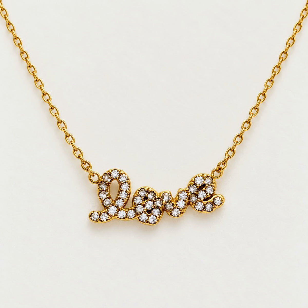 A gold chain necklace with a cursive 'love' pendant paved with small clear stones, shown on a plain light background.