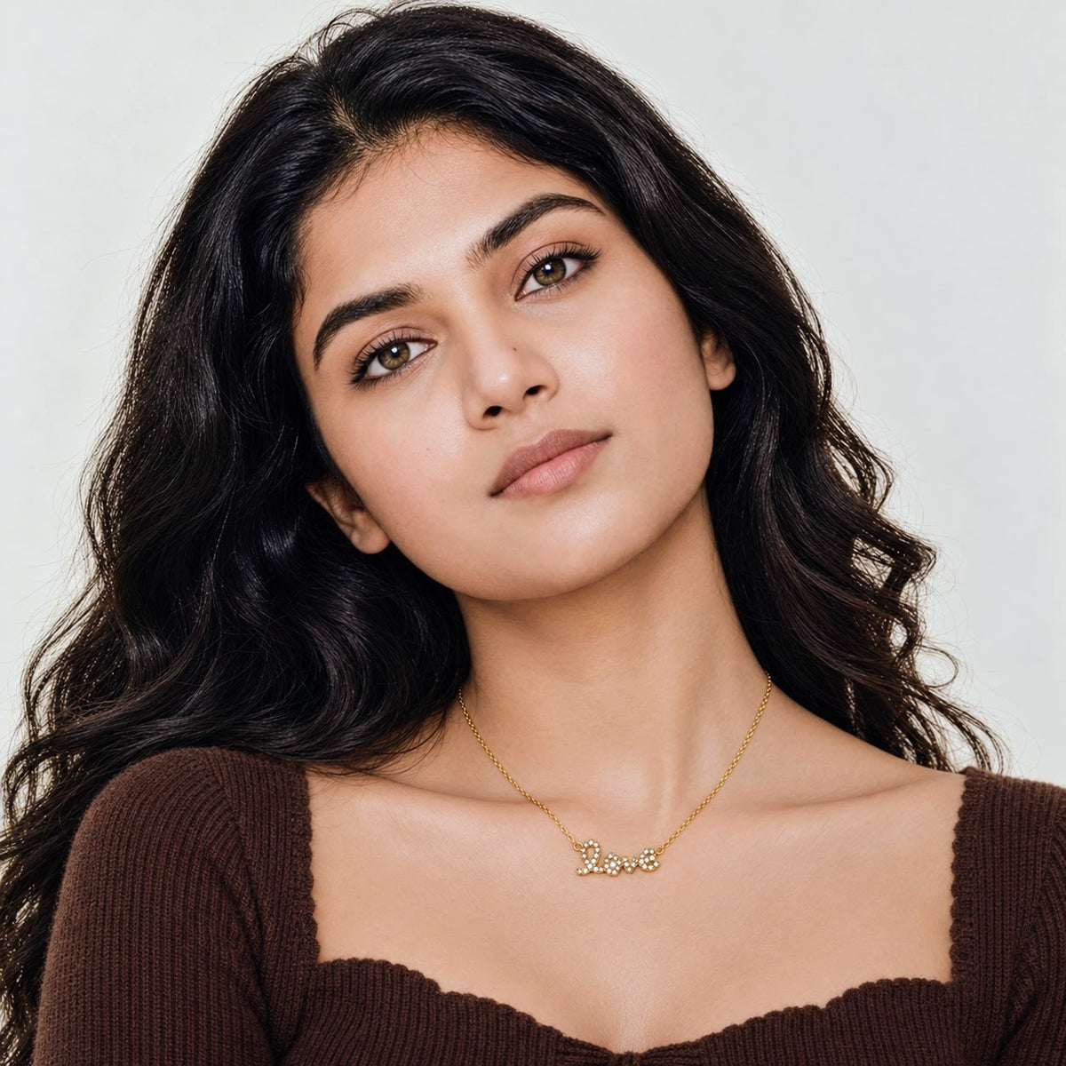 A woman with long, wavy dark hair is shown from the chest up, wearing a delicate gold necklace with a script lettering 'Love' pendant. She is wearing a brown ribbed top against a clean, light gray background.