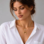 A close-up of a woman wearing two delicate gold necklaces, including a crescent moon pendant and a small round charm, styled with a white button-down shirt against a neutral gray background.
