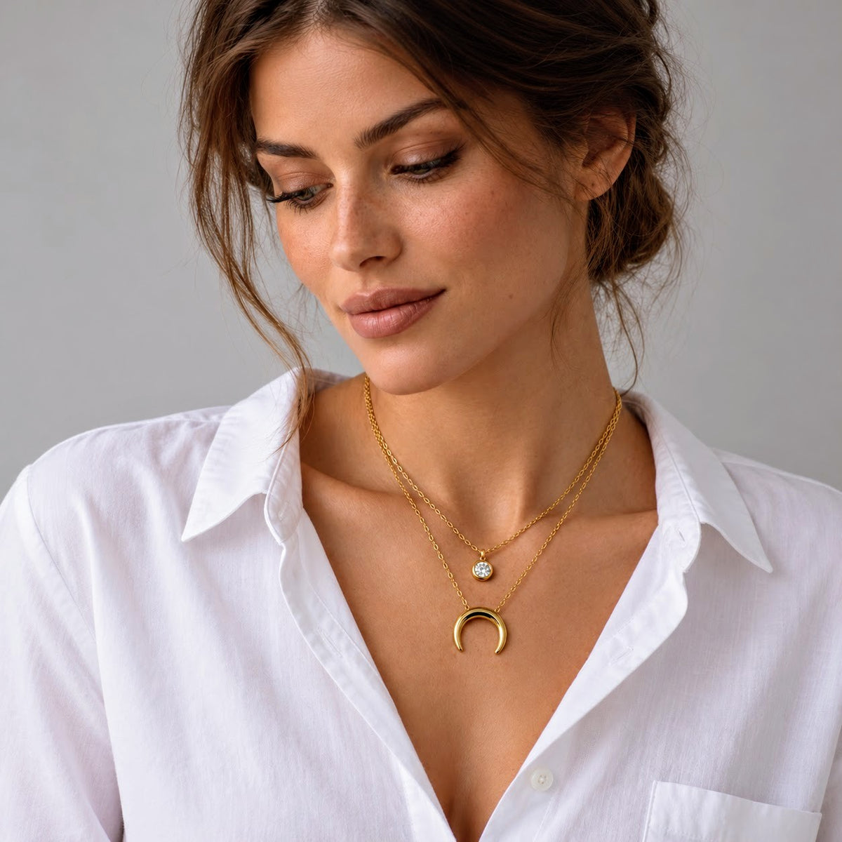 A close-up of a woman wearing two delicate gold necklaces, including a crescent moon pendant and a small round charm, styled with a white button-down shirt against a neutral gray background.