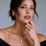 A close-up portrait of a woman wearing a gold chain necklace with a teardrop-shaped clear gemstone pendant, along with matching gold hoop earrings and a gold ring, against a soft gray background.