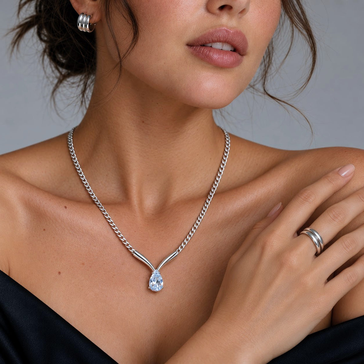 A close-up lifestyle shot of a model wearing a silver Y-shaped chain necklace with a teardrop-cut clear gemstone pendant. The necklace is displayed on her neck, with her hand resting on her shoulder against a neutral gray background.