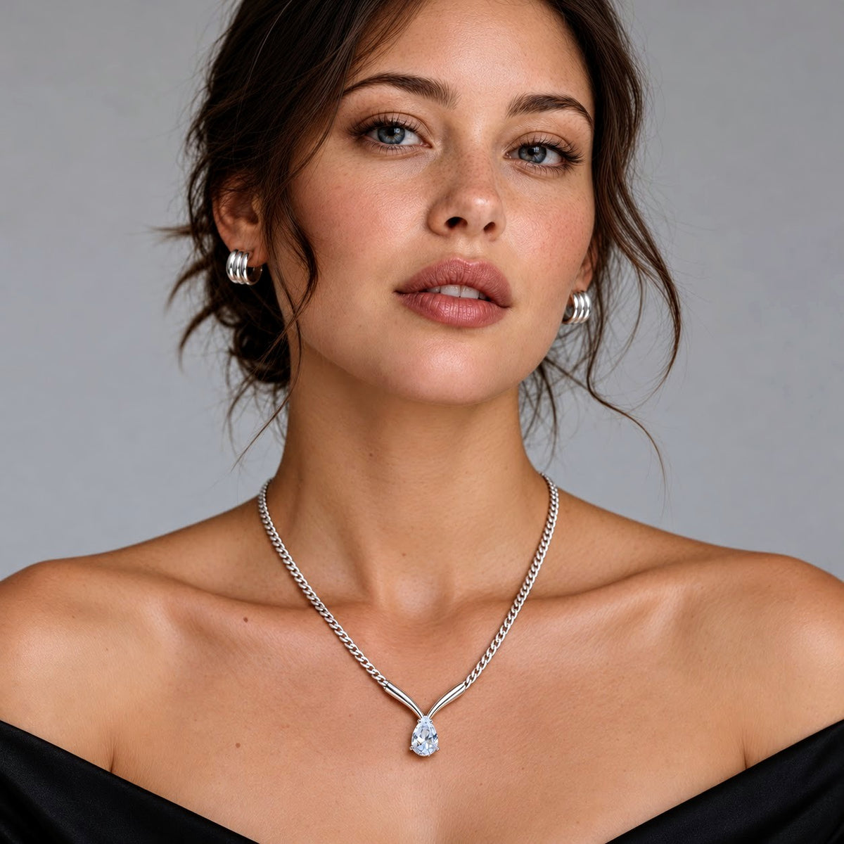 A woman wearing a silver Y-shaped chain necklace with a teardrop-shaped clear gemstone pendant, styled with matching silver hoop earrings against a neutral gray background.