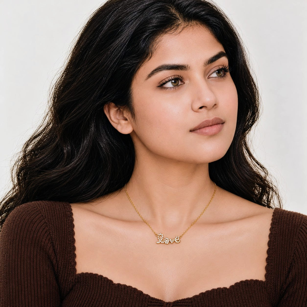 A woman with long dark hair wearing a delicate gold necklace with the word 'Love' spelled out in small letters, paired with a brown ribbed top against a light gray background.