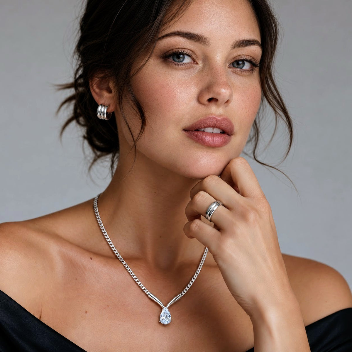 A close-up portrait of a woman wearing a silver chain necklace with a teardrop-shaped clear gemstone pendant, along with matching silver hoop earrings and a silver ring. The background is a solid neutral gray.