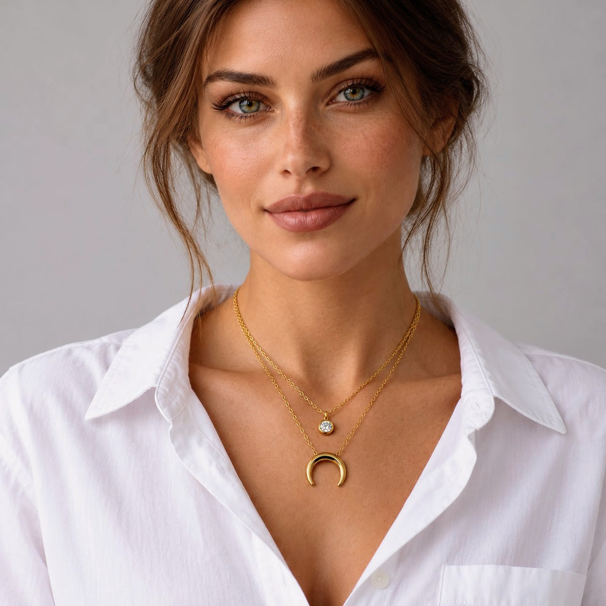 A close-up of a woman wearing a gold crescent moon pendant necklace and a delicate gold chain necklace with a small clear stone, paired with a white collared shirt against a neutral gray background.