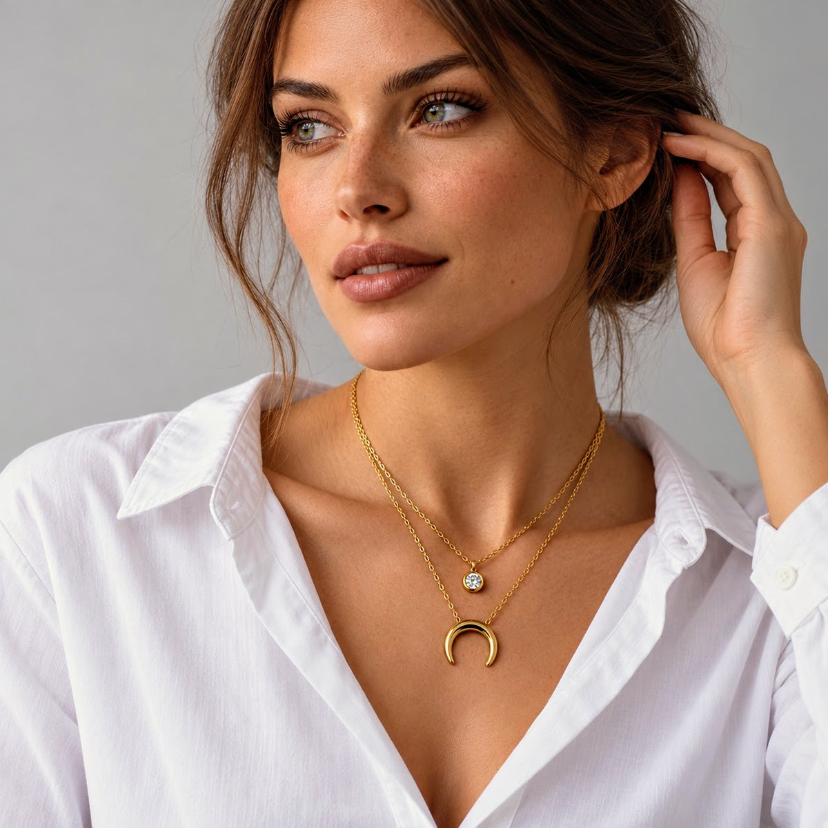 A close-up of a woman wearing a double-layered gold necklace with a crescent moon pendant and a small round stone charm, paired with a white collared shirt against a neutral gray background.