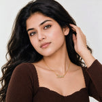 A woman with long, wavy dark hair wearing a brown ribbed knit top and a delicate gold nameplate necklace that spells 'Love'. She is posing against a clean, light off-white background.