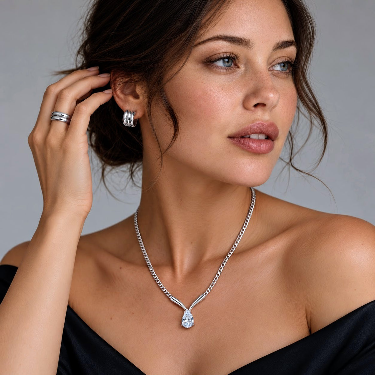 A woman wearing a silver-toned Y-shaped pendant necklace with a teardrop-cut clear gemstone and matching stud earrings, styled with a black off-the-shoulder top against a neutral gray background.