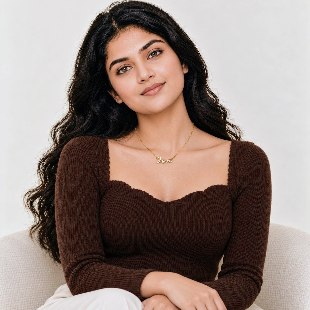A woman with long, wavy dark hair is seated on a light-colored sofa, wearing a ribbed brown long-sleeve top with a scalloped neckline and a delicate gold necklace.