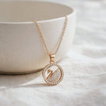 A rose gold necklace with a circular pendant featuring a swan design paved with small clear stones, draped over a white ceramic bowl on a textured white fabric background.